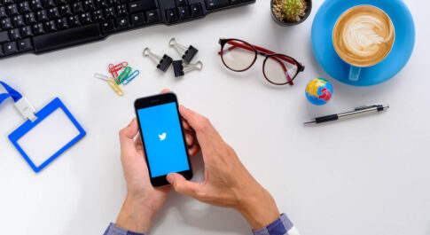 męskie dłonie trzymające telefon z logo Twittera, które opierają się na biurku pełnym innych przedmiotów, np. globus, okulary, spinacze