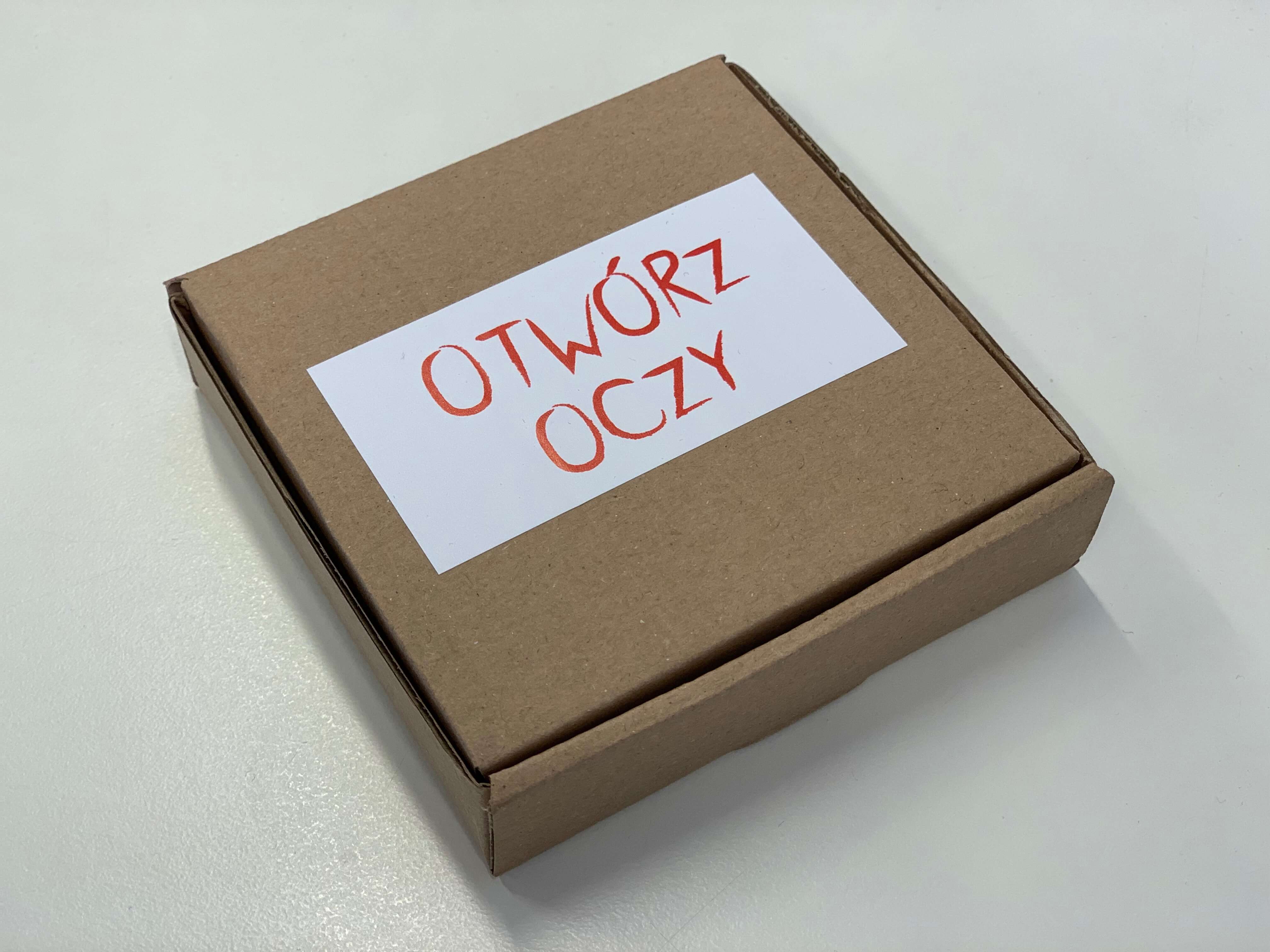 Tekturowe pudełeczko, z naklejoną białą karteczką z czerwonym napisem "Otwórz oczy"