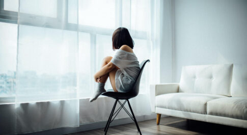 Nastolatka siedząca tyłem, z podkurczonymi nogami, na krześle. Patrzy w okno, z białą, długa firanką.
