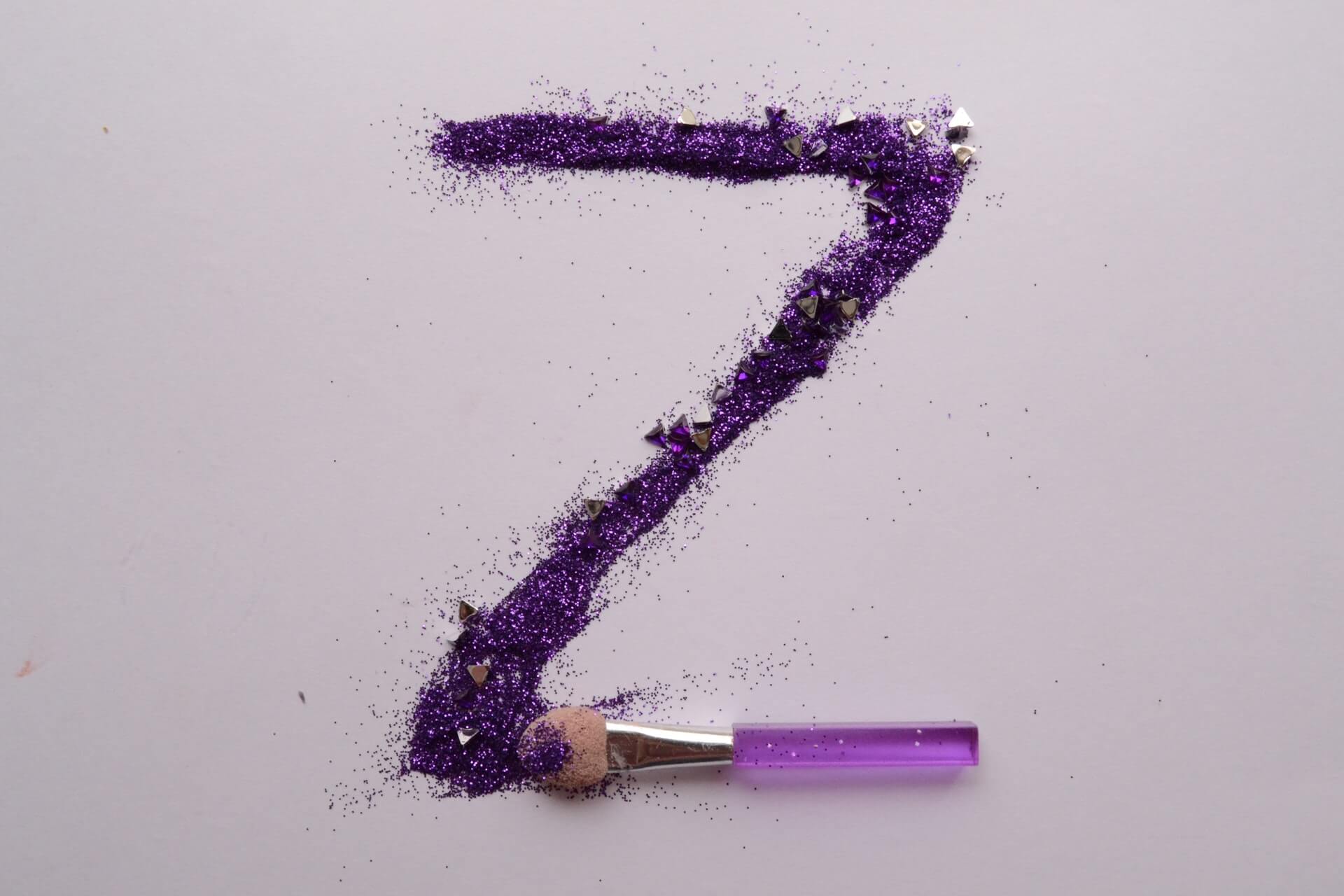 Wielkie, fioletowe "Z" napisane cieniem do powiek. Na dole litery znajduje się pacynka do makijażu. Wszystko na tle w kolorze przybrudzonego różu.