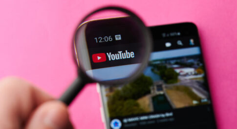Telefon z włączonym YouTube leży na ciemnym, różowym tle. Ktoś trzyma lupę, przez którą powiększone jest logo serwisu.