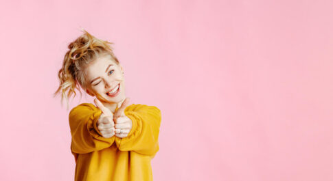 Dziewczyna pokazująca dwa kciuki, w żółtym swetrze. Stoi na różowym tle.