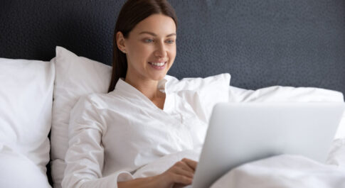 Kobieta w białej koszuli siedząca na łóżku z laptopem.