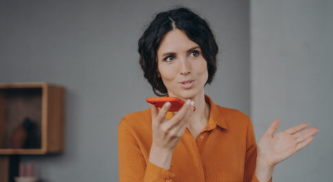 Kobieta w pomarańczowej koszuli, czarnych włosach, trzyma w prawym ręku telefon w pomarańczowej obudowie, do którego mówi. Znajduje się w domu, w którym ściany pomalowane są na szaro.