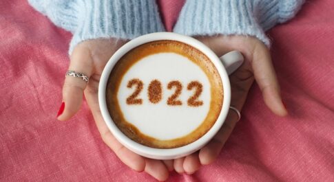 Kobiece dłonie trzymające filiżankę wypełnioną kawą z pianką. Na piance jest napis 2022.