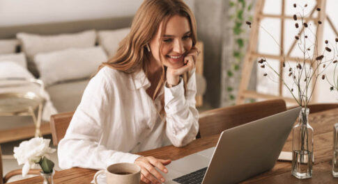 Kobieta w białej koszuli siedząca przed ekranem laptopa, w domu. Po jej prawej stronie stoi kubek z kawą.