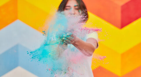 Kobieta dmuchająca w kolorowy pyłek. Na tle kolorowej ściany.