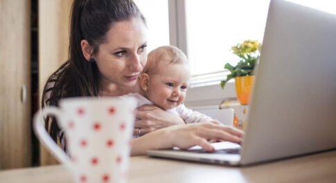 Matka trzymająca dziecko na kolanach, siedząca przed komputerem. Na pierwszym planie znajduje się też biały kubek, w czerwone kropki.