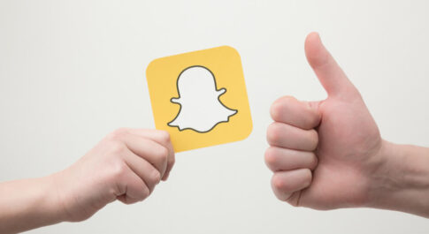 Na biały tle, jedna ręka trzyma kartkę z logo Snapchata, druga pokazuje kciuk do góry.- Snapchat