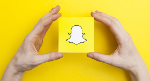 Na żółtym tle, dwie dłonie trzymają klocek z logo Snapchata.