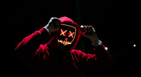 Na czarnym tle, postać ubrana w czerwoną bluzę, z kapturem na głowie. Ma maskę, z ledowymi oczami (x) oraz ledowymi ustami, przypominającymi zaszyte wargi.