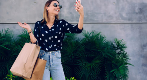 Kobieta robiąca sobie selfie, w drugim ręku trzyma torby zakupowe. Stoi na tle betonowej ściany.