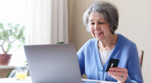 Starsza kobieta robiąca zakupy online. Siedzi przed laptopem i trzyma kartę kredytować w ręku.