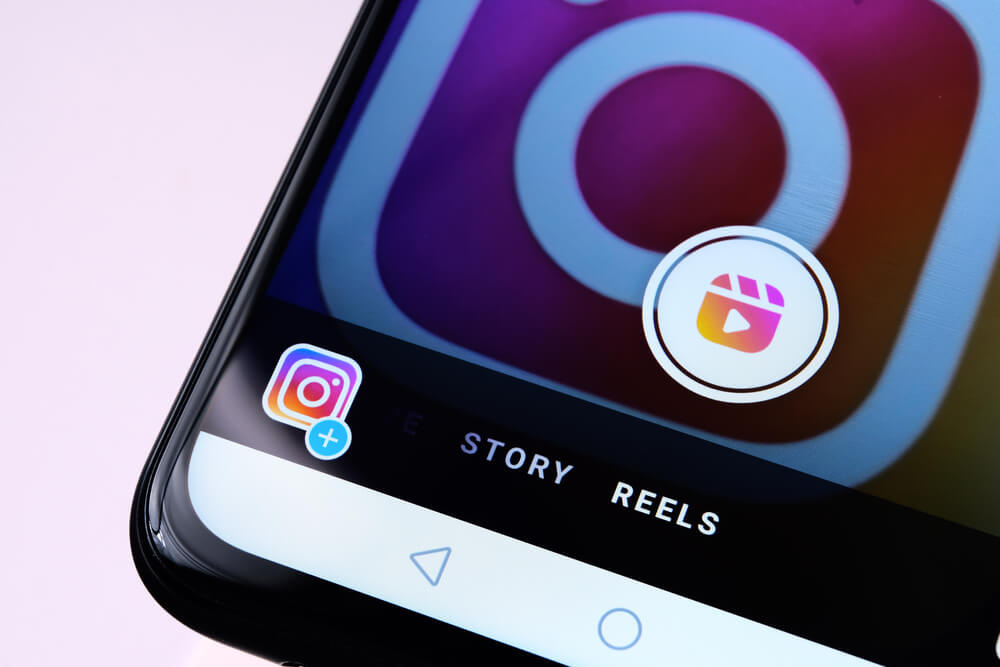 Telefon leżący na różowym tle. Widać ikonę Instagram oraz zakładki Story i Reels.
