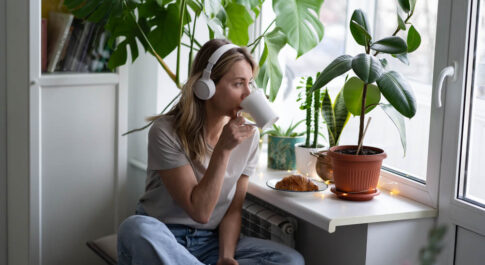 Kobieta ze słuchawkami na uszach, siedzi po turecku na stołku. Patrzy w okno, pije coś z białego kubka. Dookoła niej dużo kwiatów zielonych, doniczkowych.